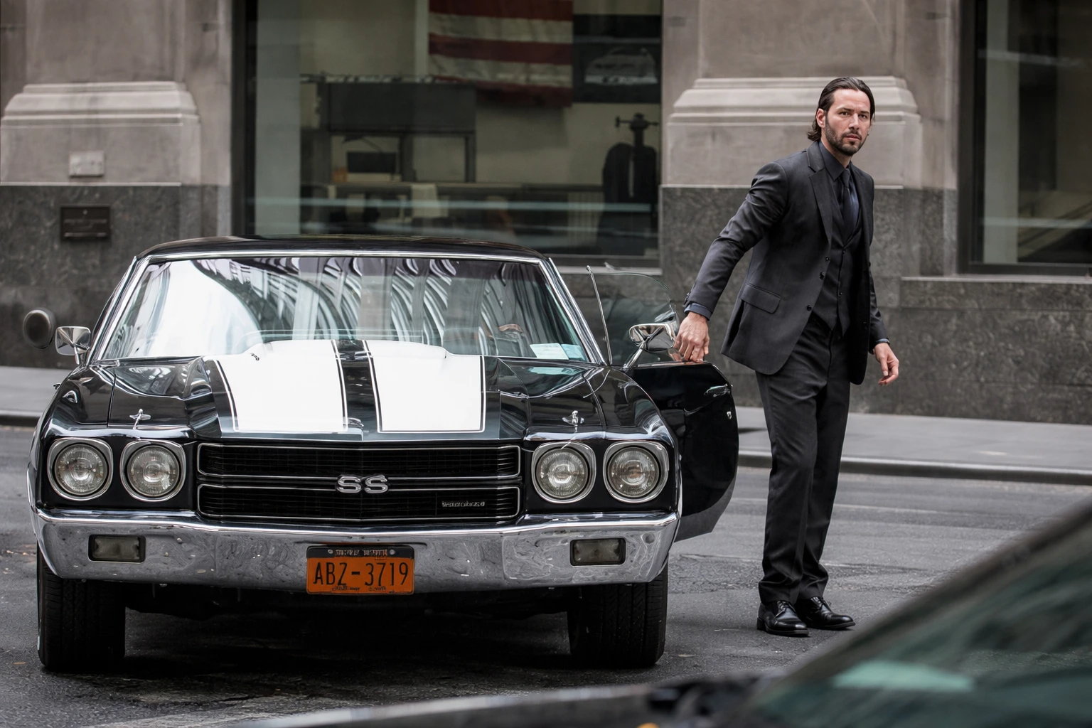John Wick en costume noir sortant d’une Chevrolet Chevelle SS classique noire avec bandes blanches